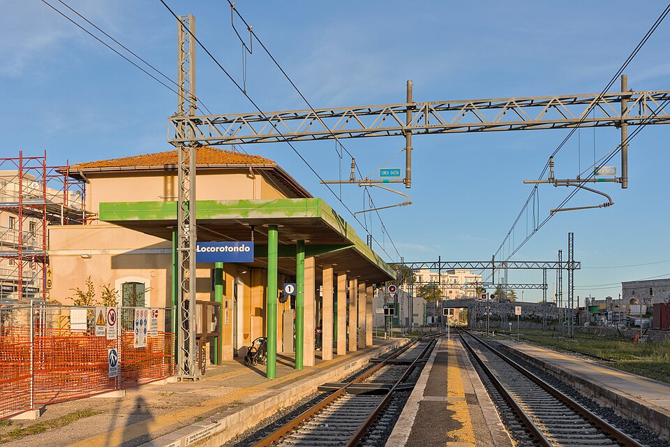 Gare de Locorotondo : bâtiment voyageurs et voie 1 sous l’auvent vert, avec la ligne aérienne et une clôture de chantier près du bâtiment